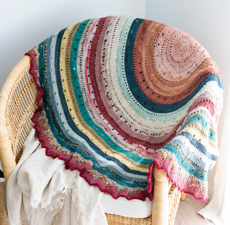 A large circular knitted throw with colourful stripes, eyelet lace and a chevron lace border